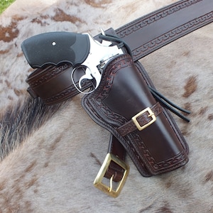 Op de afbeelding: Een bruine leren holster met een zilveren revolver en een bruine leren riem met ingewikkelde stiksels. De holster en riem bevinden zich op een bruin en wit dierenhuid.