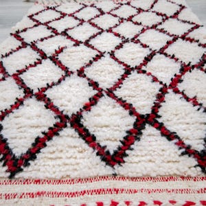 May include: A white rug with a diamond pattern in red and black. The rug has a textured, plush surface and a red border with a zigzag design. The rug is a decorative home accent.