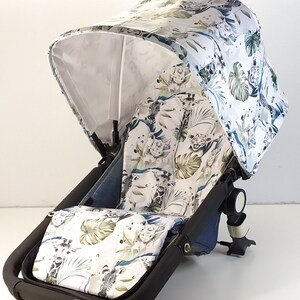 May include: A stroller canopy and seat cover featuring a jungle-themed pattern. The white fabric is adorned with illustrations of animals, including giraffes and lions, along with green and blue foliage. The stroller frame is black.