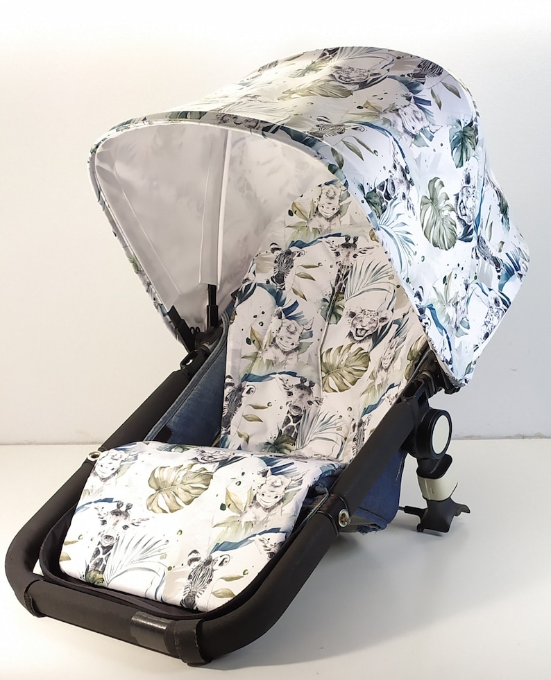 May include: A stroller canopy and seat cover featuring a jungle-themed pattern. The white fabric is adorned with illustrations of animals, including giraffes and lions, along with green and blue foliage. The stroller frame is black.