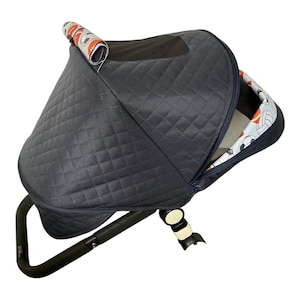 May include: A navy blue quilted stroller canopy with a black handle. The canopy has a patterned interior with soccer balls and orange accents. The stroller's design is modern and functional.