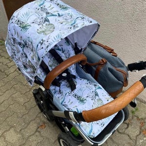 May include: A baby stroller with a white canopy and seat featuring a jungle-themed pattern with green leaves and animal prints. The stroller has brown leather handles and a matching diaper bag.