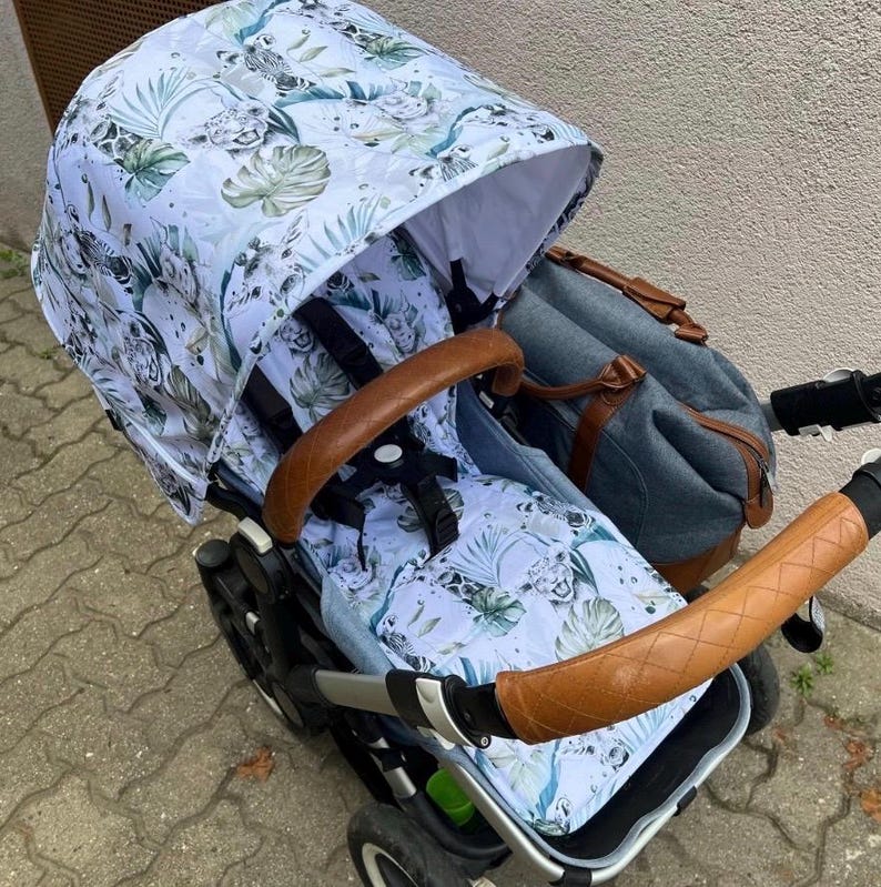 May include: A baby stroller with a white canopy and seat featuring a jungle-themed pattern with green leaves and animal prints. The stroller has brown leather handles and a matching diaper bag.