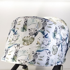 May include: A white stroller canopy featuring a jungle-themed pattern. The design includes illustrations of giraffes, zebras, and lions amidst green tropical leaves. The canopy is curved and appears to be made of fabric.