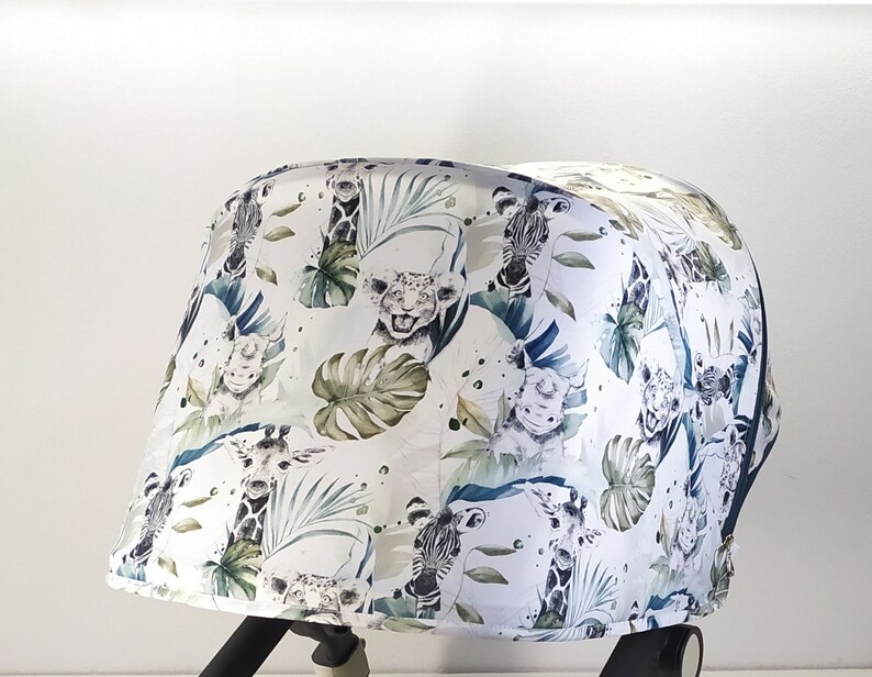 May include: A white stroller canopy featuring a jungle-themed pattern. The design includes illustrations of giraffes, zebras, and lions amidst green tropical leaves. The canopy is curved and appears to be made of fabric.