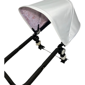 Pode incluir: Uma capota de carrinho de bebé branca com um padrão floral no interior. A capota está presa às pegas e à estrutura preta do carrinho. O design proporciona sombra e proteção solar.