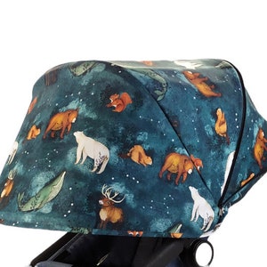 Könnte beinhalten: Ein Kinderwagenverdeck mit dunkelblaugrünem Hintergrund, der Illustrationen von Bären, Hirschen, Walen und Eichhörnchen zeigt. Das Verdeck hat ein Sternenhimmel-Design mit Sternbildern und weißen Akzenten. Es ist an einem schwarzen Kinderwagengestell befestigt.