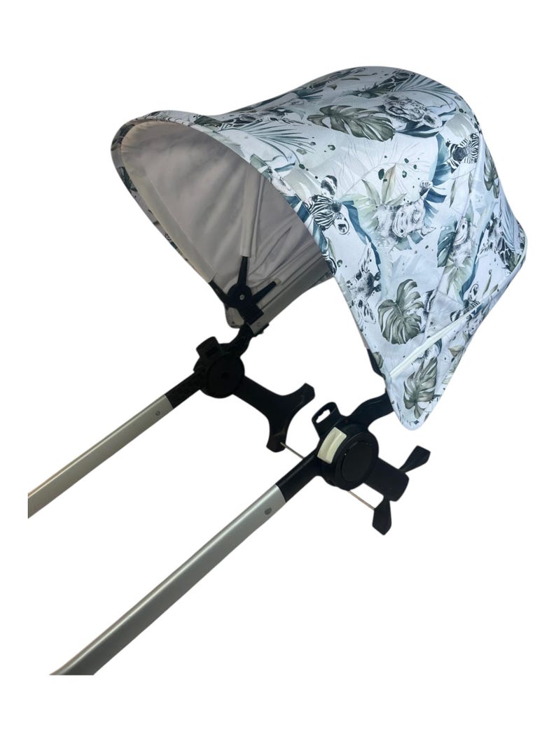 May include: A stroller canopy with a white background and a jungle-themed pattern featuring green leaves and animal prints. The canopy is attached to silver metal poles and black joints.