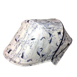 May include: A white and blue patterned baby stroller sunshade. The fabric features a crane and bamboo design. The cranes are depicted in flight and standing, with red accents on their heads. The sunshade has a dark blue trim.