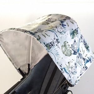 May include: A stroller canopy with a white background and a jungle-themed pattern. The pattern includes green leaves, giraffes, and other animals. The canopy is attached to a black stroller frame.