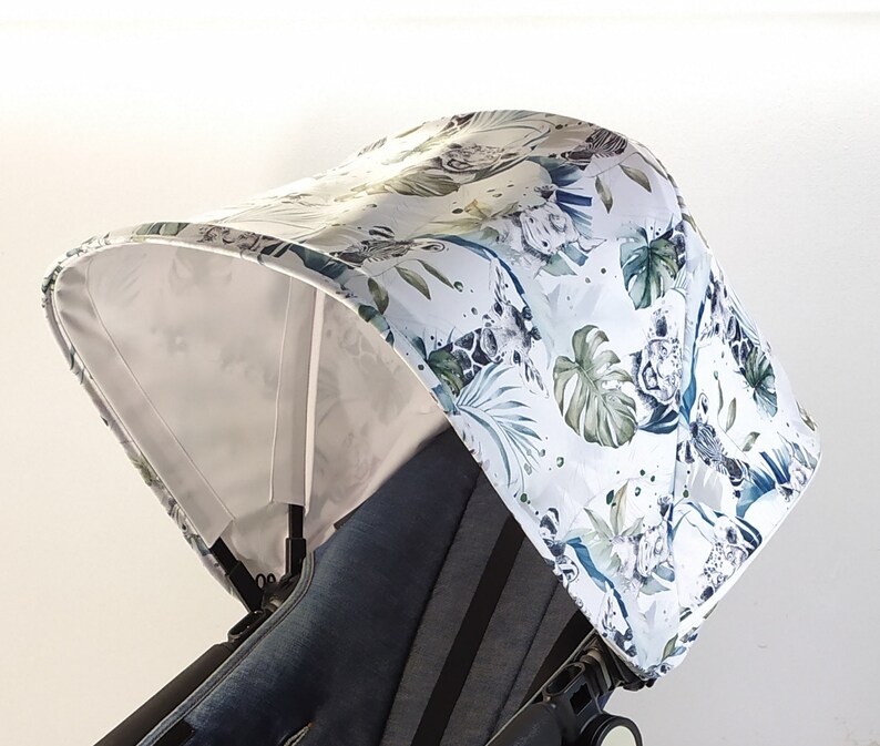 May include: A stroller canopy with a white background and a jungle-themed pattern. The pattern includes green leaves, giraffes, and other animals. The canopy is attached to a black stroller frame.