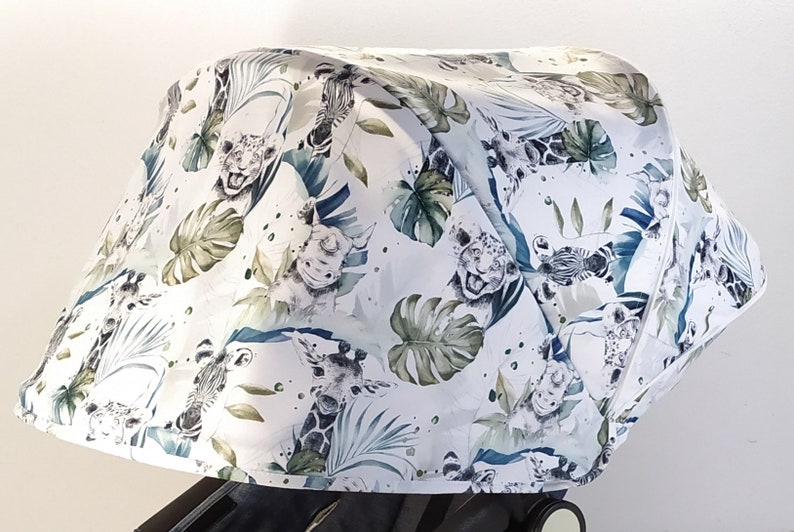 May include: A white stroller canopy featuring a jungle-themed pattern. The design includes illustrations of giraffes, zebras, tigers, and tropical foliage in shades of green and blue. The canopy is attached to a black stroller frame.