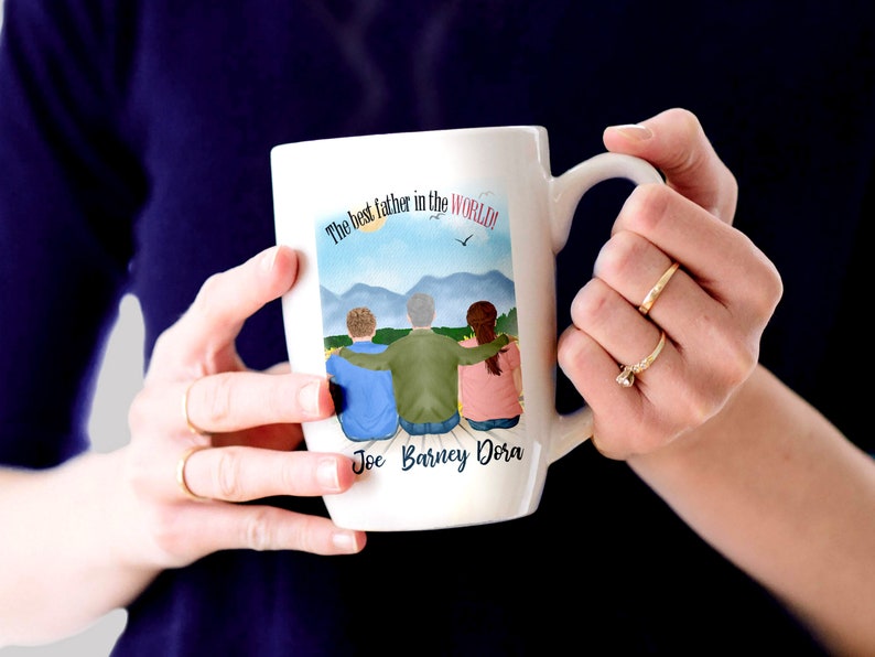 May include: White ceramic mug featuring a colourful illustration of three people standing together with the text "The best father in the world!" and the names "Joe Barney Dora" printed below the illustration.