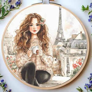 以下が含まれることがあります： 木製のフレームに入ったクロスステッチ刺繍。長い茶色の髪の女性がコーヒーカップを持ち、背景にエッフェル塔が描かれています。女性はベージュのセーターと黒いパンツを着用しています。作品はラベンダーに囲まれています。