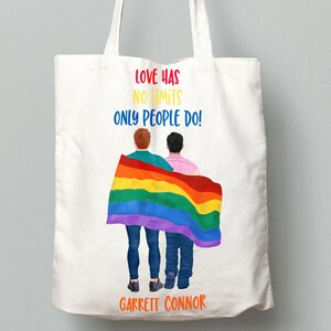 May include: White tote bag with a rainbow flag and two people standing in front of it. The text on the bag reads "Love has no limits only people do!" and "Garrett Connor".