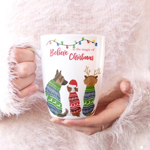 May include: White ceramic mug with a Christmas-themed design. The mug features three dogs wearing festive sweaters and the text "Believe in the magic of Christmas".