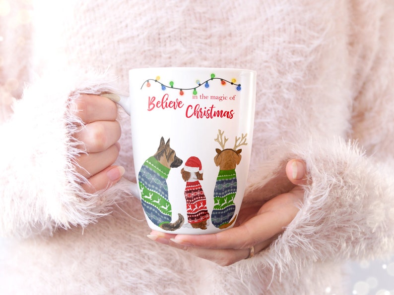 May include: White ceramic mug with a Christmas-themed design. The mug features three dogs wearing festive sweaters and the text "Believe in the magic of Christmas".