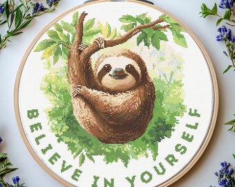 May include: A cross-stitch embroidery hoop featuring a brown sloth hanging from a tree branch, surrounded by green leaves. The text "BELIEVE IN YOURSELF" is stitched in green around the sloth. The hoop is made of wood.
