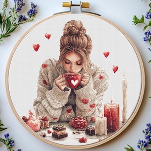 May include: A cross-stitch pattern of a woman in a white sweater with a red heart on her mug, surrounded by red hearts and candles.