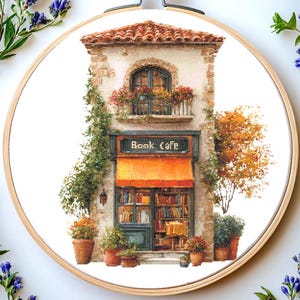 Pode incluir: Bordado em ponto cruz de um pitoresco café-livraria. O edifício apresenta um telhado de telhas vermelhas, um toldo laranja e uma placa que diz "Book Cafe". A fachada é adornada com flores e vegetação, e o interior da loja é cheio de livros.