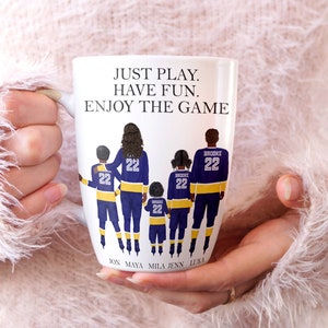 May include: White ceramic coffee mug with a graphic of a family of five wearing hockey jerseys. The text "JUST PLAY. HAVE FUN. ENJOY THE GAME" is printed on the mug. The names "JON, MAYA, MILA, JENN, LUKA" are printed below the graphic.