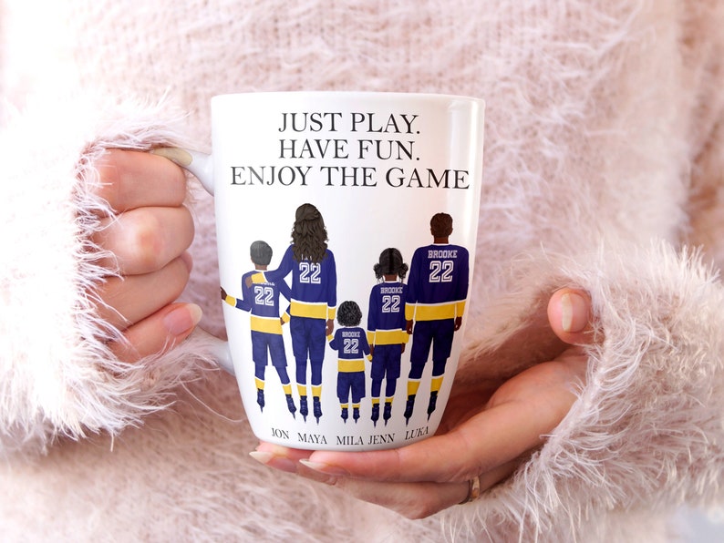 May include: White ceramic coffee mug with a graphic of a family of five wearing hockey jerseys. The text "JUST PLAY. HAVE FUN. ENJOY THE GAME" is printed on the mug. The names "JON, MAYA, MILA, JENN, LUKA" are printed below the graphic.