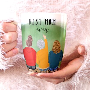 May include: White ceramic mug with a green background and three women standing on a wooden dock. The text "BEST MOM ever" is printed on the mug. The women are wearing red, yellow, and blue sweaters. Their names are printed below them: "DONNA, AUDRA, SARAH".