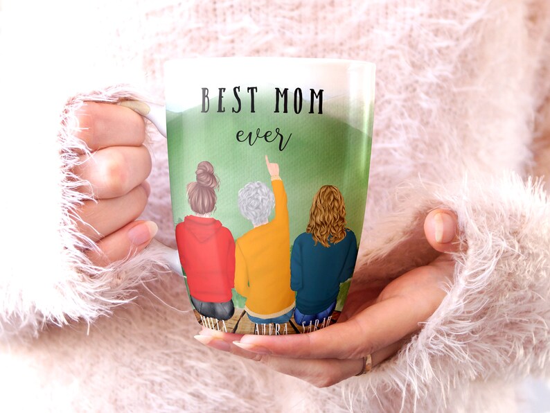 May include: White ceramic mug with a green background and three women standing on a wooden dock. The text "BEST MOM ever" is printed on the mug. The women are wearing red, yellow, and blue sweaters. Their names are printed below them: "DONNA, AUDRA, SARAH".