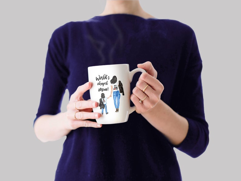 Op de afbeelding: Een witte keramische mok met een zwarte handgreep. De mok heeft een aquarel-illustratie van een moeder en kind die hand in hand lopen. De tekst op de mok is "World's okayest mom!"