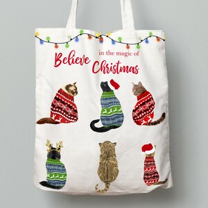 May include: White canvas tote bag with the text "Believe in the magic of Christmas" and five illustrations of cats wearing Christmas sweaters.