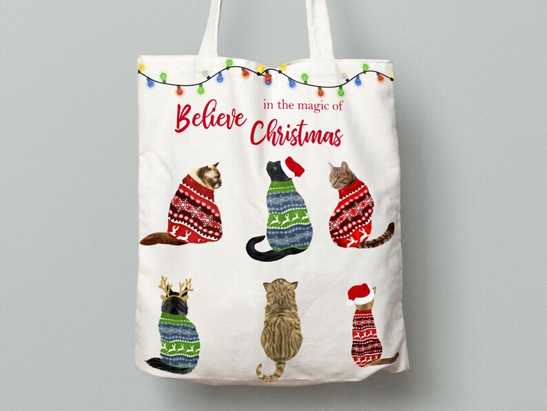 May include: White canvas tote bag with the text "Believe in the magic of Christmas" and five illustrations of cats wearing Christmas sweaters.