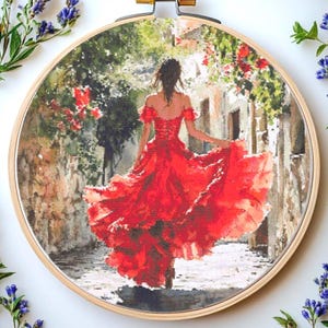 May include: A cross-stitch embroidery in a wooden hoop shows a woman in a flowing red dress walking down a cobbled street. The scene is framed by a stone wall and greenery, with red flowers adding pops of colour.