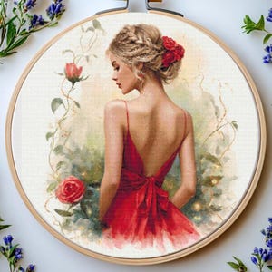 Pode incluir: Bordado em ponto cruz num bastidor de madeira com uma mulher num vestido vermelho com as costas abertas e um laço vermelho. A mulher tem cabelo loiro e rosas vermelhas. O fundo tem um efeito suave e desfocado com elementos florais.