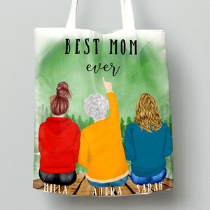 May include: A white tote bag with a green watercolor background and the text "BEST MOM ever". Three women are pictured on the bag, with their backs to the viewer, sitting on a wooden dock. The women are wearing red, yellow, and blue sweatshirts. The text "MILLA", "AUDRA", and "SARAH" are written below the women.