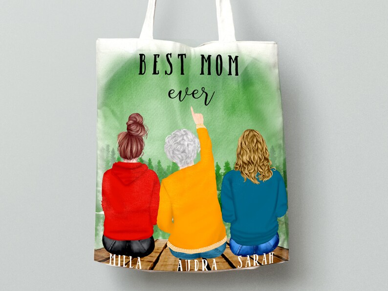 May include: A white tote bag with a green watercolor background and the text "BEST MOM ever". Three women are pictured on the bag, with their backs to the viewer, sitting on a wooden dock. The women are wearing red, yellow, and blue sweatshirts. The text "MILLA", "AUDRA", and "SARAH" are written below the women.