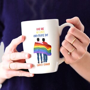 May include: White ceramic mug with a rainbow flag and two people holding hands. The text on the mug reads "Love has no limits only people do!" and "Garrett Connor".