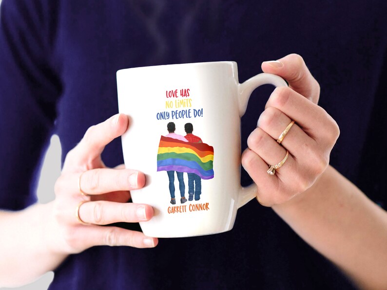 May include: White ceramic mug with a rainbow flag and two people holding hands. The text on the mug reads "Love has no limits only people do!" and "Garrett Connor".