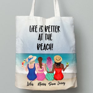 May include: A white tote bag with the text "Life is better at the beach!" printed on it. The bag features a colourful illustration of four women in swimsuits and hats on a beach with drinks in their hands. The names "Lola, Nina, Sam, Jenny" are printed below the illustration.