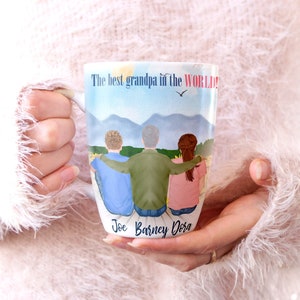 May include: White ceramic mug featuring a colourful illustration of three people standing together in front of a mountain range. The text "The best grandpa in the WORLD!" is printed at the top of the mug. The names "Joe, Barney, Dora" are printed at the bottom of the mug.