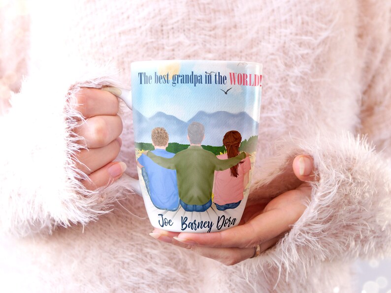 May include: White ceramic mug featuring a colourful illustration of three people standing together in front of a mountain range. The text "The best grandpa in the WORLD!" is printed at the top of the mug. The names "Joe, Barney, Dora" are printed at the bottom of the mug.