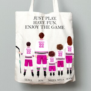 Op de afbeelding: Een witte tote bag met een roze en zwart hockeytrui-ontwerp. De tekst "JUST PLAY. HAVE FUN. ENJOY THE GAME" is op de tas gedrukt. De namen Luka, Jon, Maya, Mila en Jenn zijn onder de truien gedrukt.