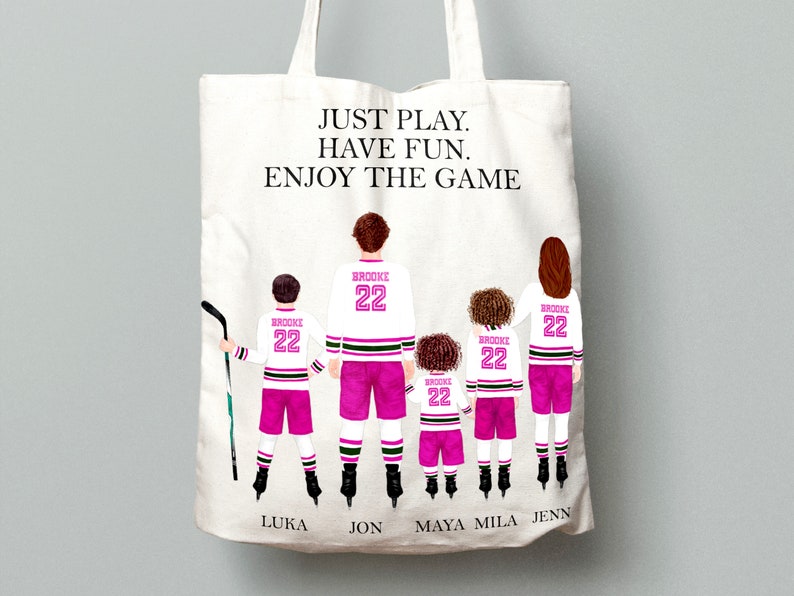 Op de afbeelding: Een witte tote bag met een roze en zwart hockeytrui-ontwerp. De tekst "JUST PLAY. HAVE FUN. ENJOY THE GAME" is op de tas gedrukt. De namen Luka, Jon, Maya, Mila en Jenn zijn onder de truien gedrukt.