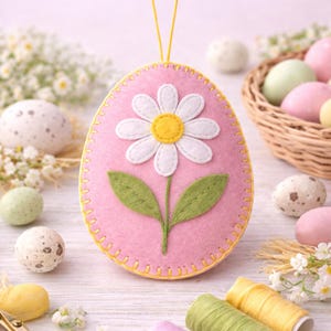Op de afbeelding: Een roze vilten paasei-ornament met een witte madelief en groene bladeren. Het ei is omrand met gele stiksels en heeft een geel touwtje om op te hangen. Decoratieve paaseieren en een mandje staan op de achtergrond.
