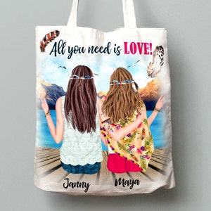 May include: White tote bag with a colorful illustration of two women with long hair and floral headbands sitting on a dock. The text "All you need is LOVE!" is printed in pink and black on the bag. The names "Janny" and "Maya" are printed at the bottom of the bag.