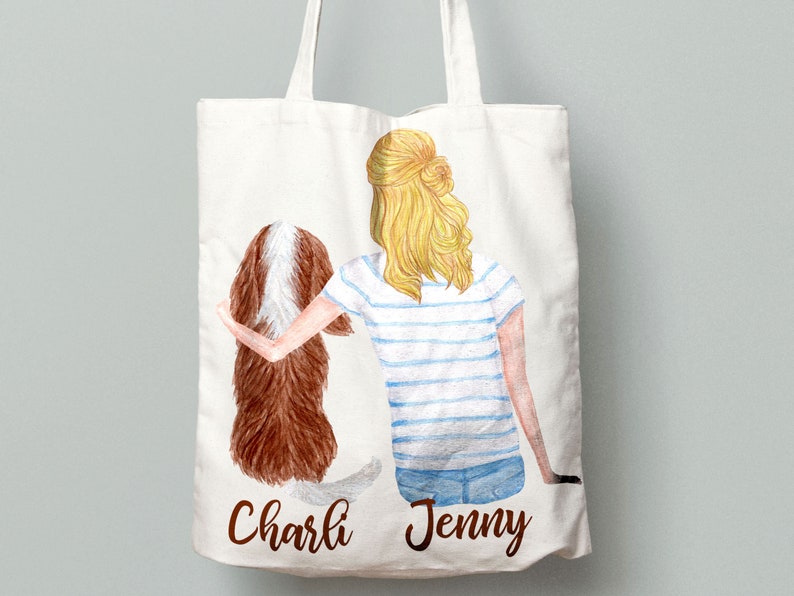May include: A white tote bag with a watercolor illustration of a woman with blonde hair and a brown and white dog. The woman is wearing a blue and white striped shirt and blue jeans. The text "Charli" and "Jenny" is printed on the bag.