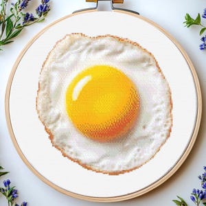 May include: A cross-stitch embroidery of a fried egg, with a vibrant yellow yolk and a white, lightly browned egg white. The embroidery is displayed in a wooden hoop, with floral decorations in the background.