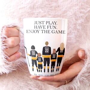 Op de afbeelding: Een witte keramische mok met een ontwerp van een hockey-speler familie in zwart en geel. De tekst "JUST PLAY. HAVE FUN. ENJOY THE GAME" is op de mok gedrukt. De namen "JON, MAYA, LUKA, MILA, JENN" staan ​​onderaan de mok.