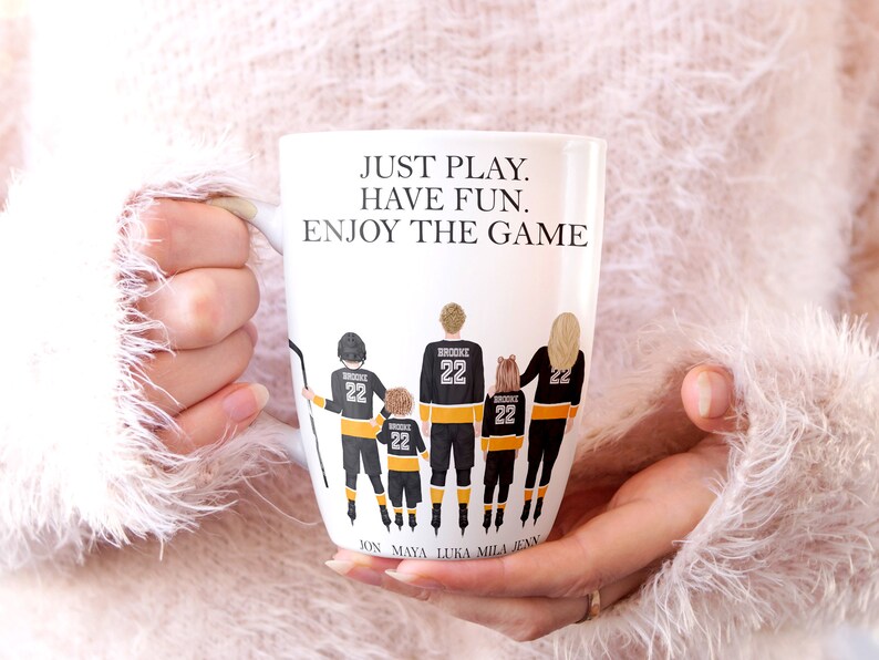 Op de afbeelding: Een witte keramische mok met een ontwerp van een hockey-speler familie in zwart en geel. De tekst "JUST PLAY. HAVE FUN. ENJOY THE GAME" is op de mok gedrukt. De namen "JON, MAYA, LUKA, MILA, JENN" staan ​​onderaan de mok.