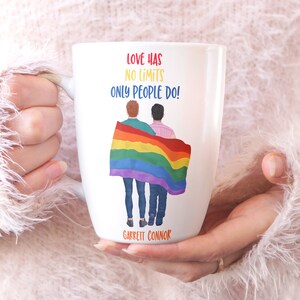 May include: White ceramic mug with a rainbow flag and two people standing in front of it. The text on the mug reads "Love has no limits only people do!" and "Garrett Connor".