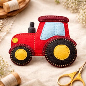 Op de afbeelding: Een handgemaakte vilten tractor in rood, met zwarte wielen en een gele kern. De tractor heeft een lichtblauw raam en een donkerrood dak. Het ontwerp is gestikt met gele draad en een gele koplamp is zichtbaar.